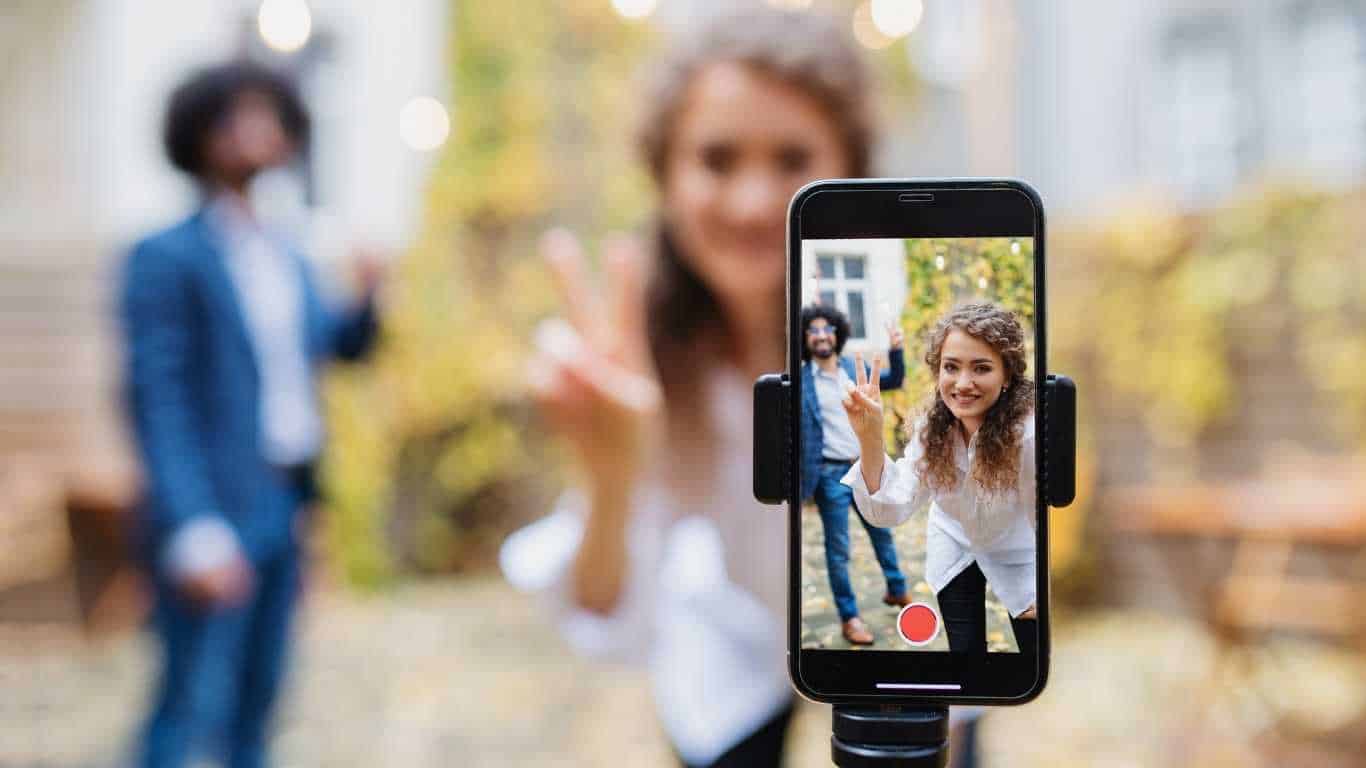 A woman taking a selfie video with two people waving in the background.