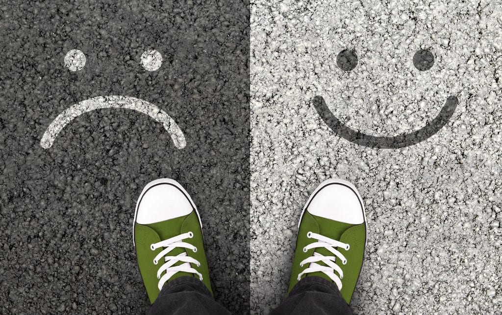 someone showing his feet in a pair of green shoes, one leg standing on a divided asphalt with smiley face on the right, and frowning face on the left