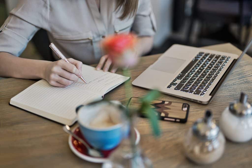 Freelance copywriter writing notes in a notebook beside a laptop and coffee on a desk