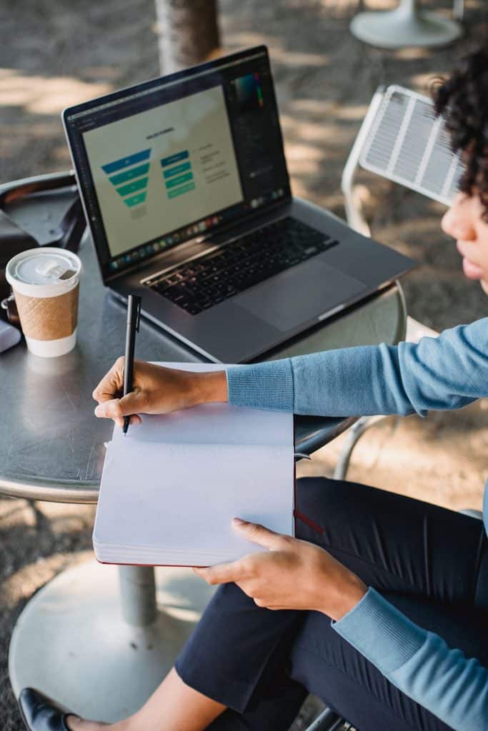 Freelancer sketching a funnel diagram in a notebook beside a laptop at a cafe