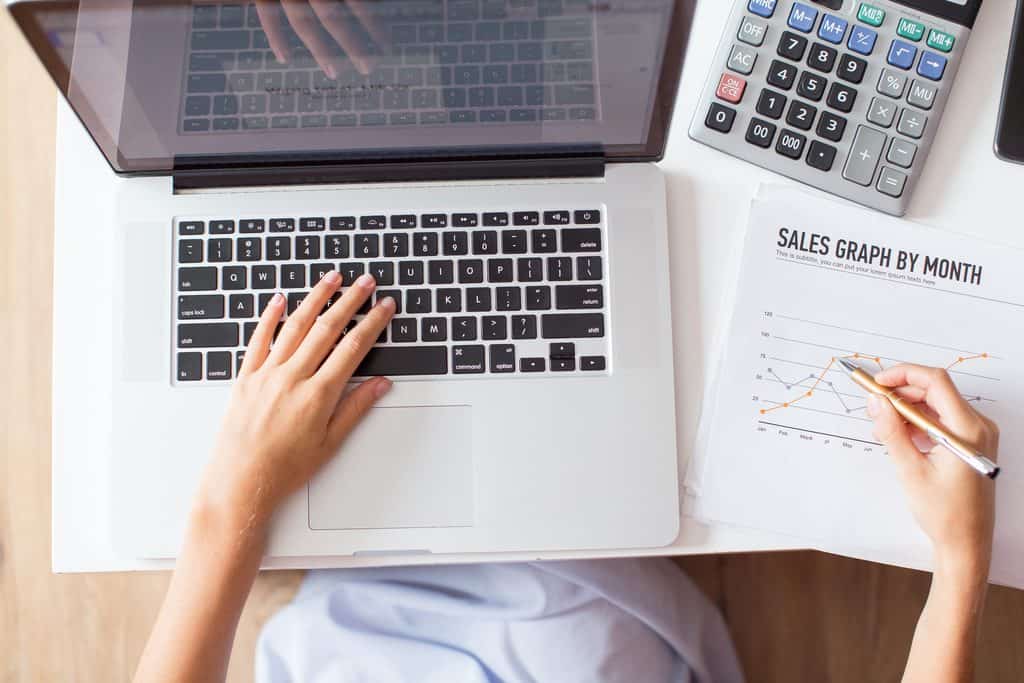 Overhead view of hands typing on a laptop beside a printed sales graph and calculator