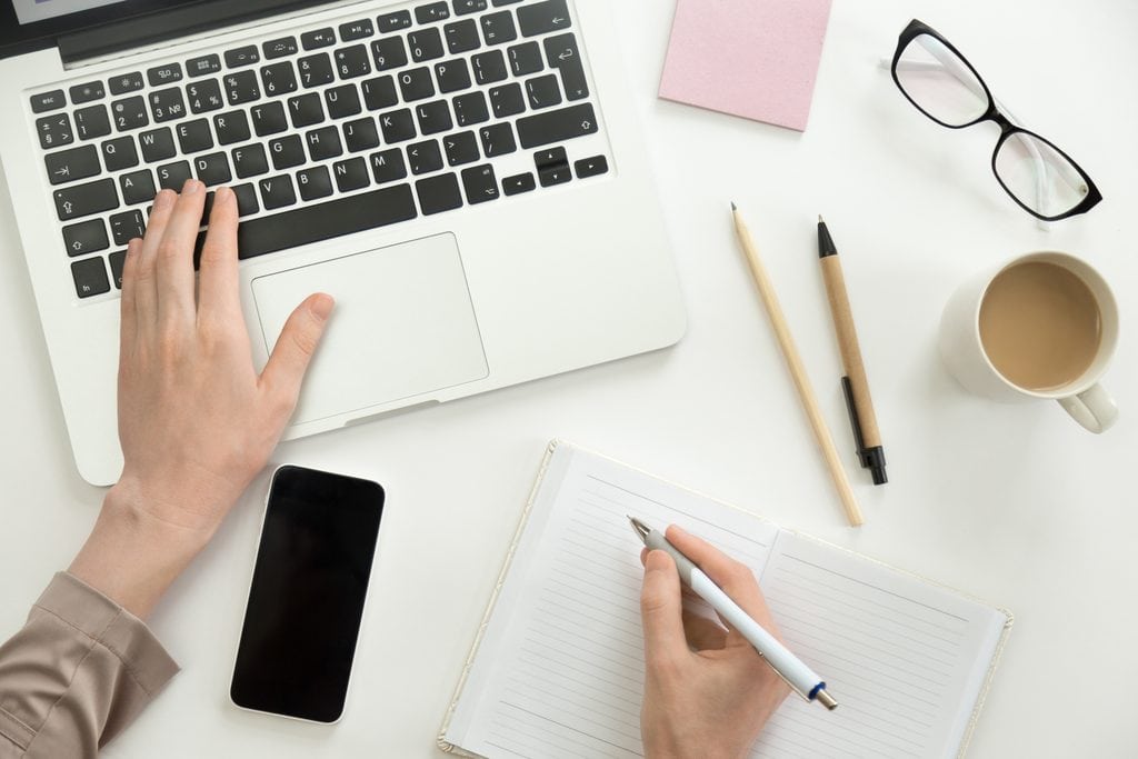 Overhead view of hands typing on a laptop while noting ideas in a notebook