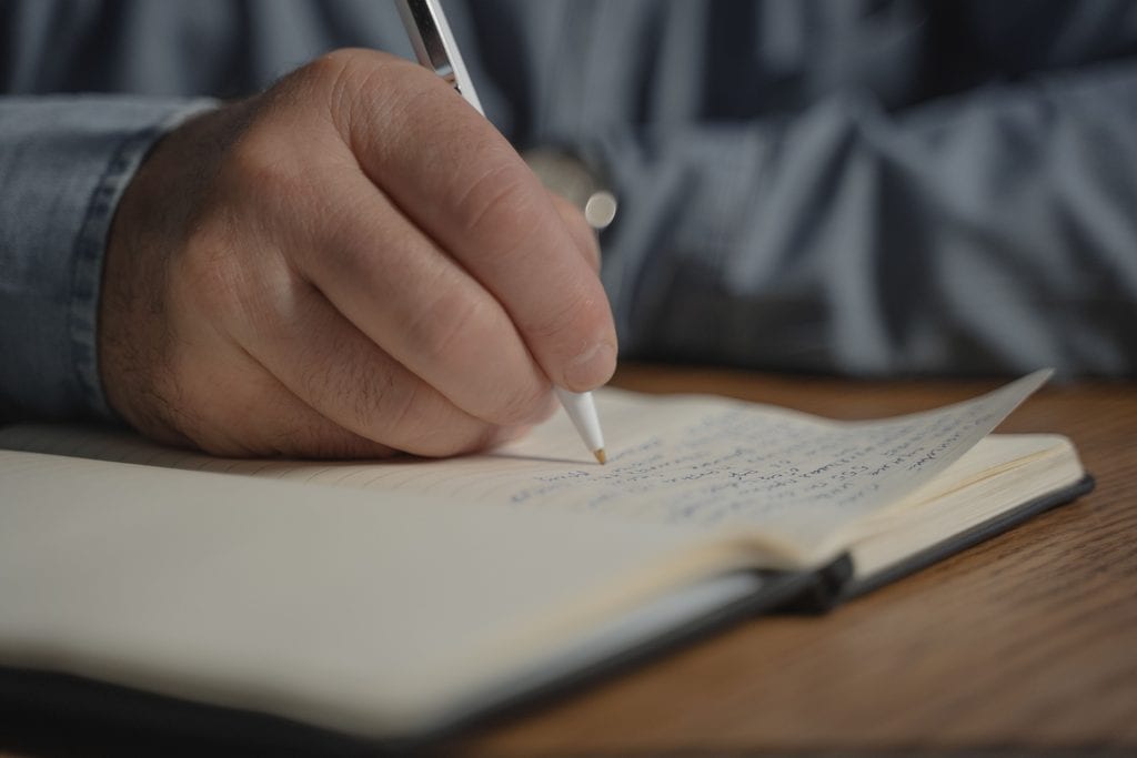 Close-up of a hand writing notes in a notebook with a pen on a wooden desk