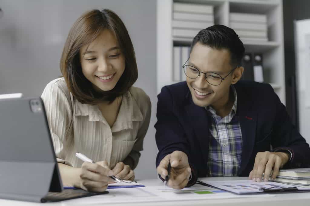 Two smiling coworkers reviewing colorful bar charts and reports on a tablet and paper documents at a desk.