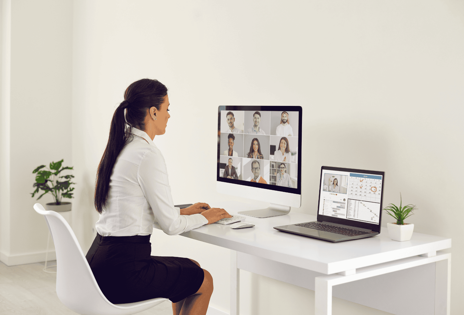 Woman working remotely at a desk with a laptop and desktop monitor showing a virtual meeting and digital work tools.