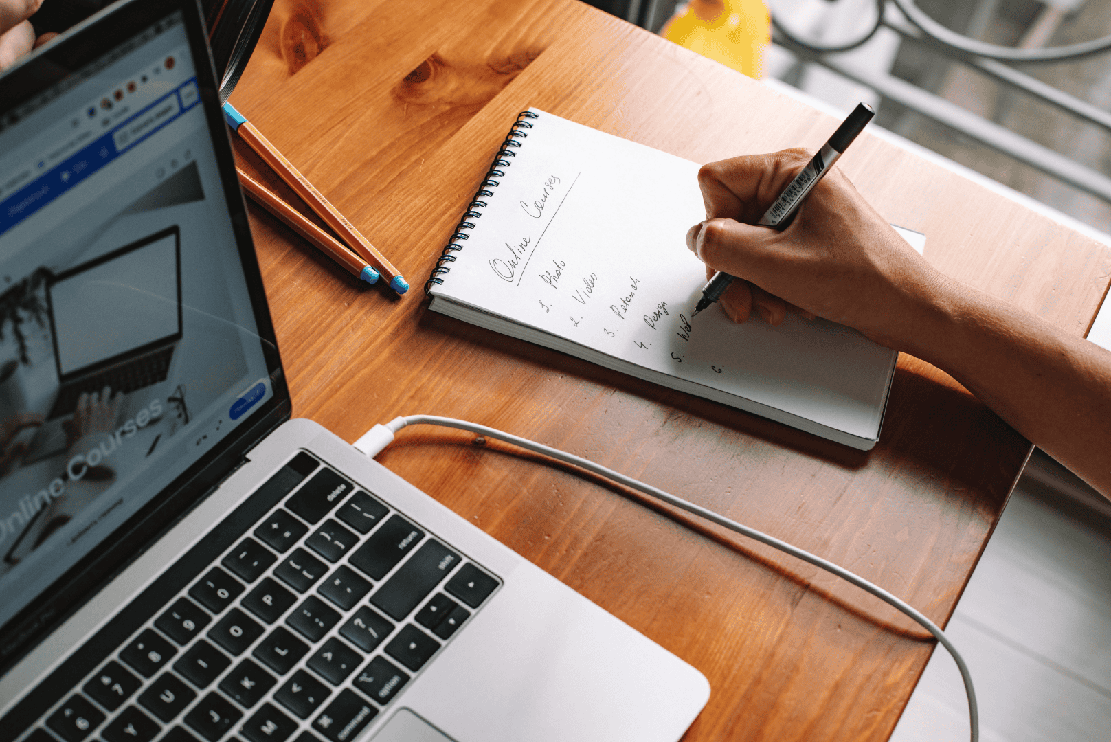 A person taking detailed notes in a notebook while researching a freelancing course for beginners on their laptop.