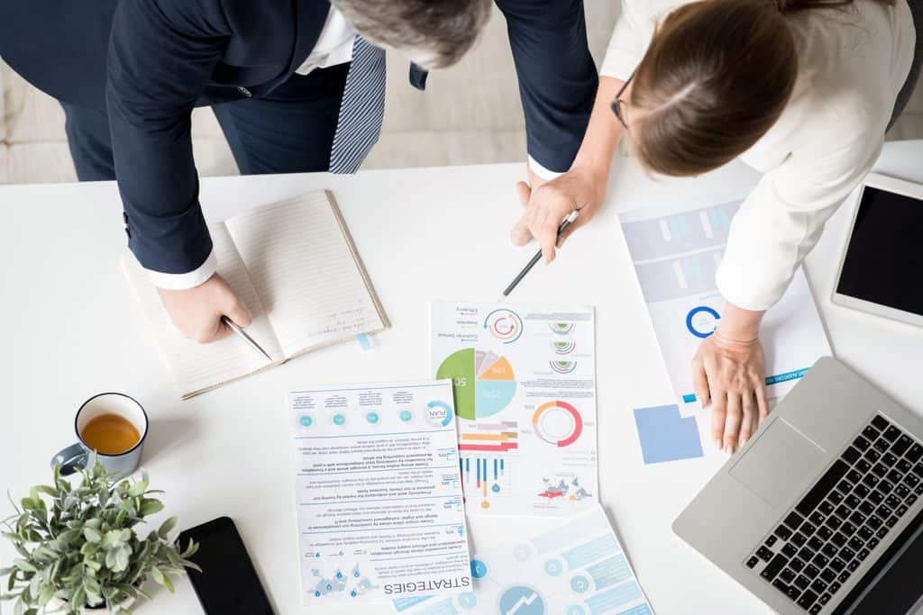 Overhead view of two colleagues pointing at printed pie charts and a strategy document on a desk with a laptop, notebook, and coffee cup nearby.