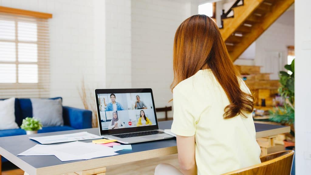 Woman viewed from behind sitting at a desk covered in papers and sticky notes, on a video call with four colleagues on her laptop.