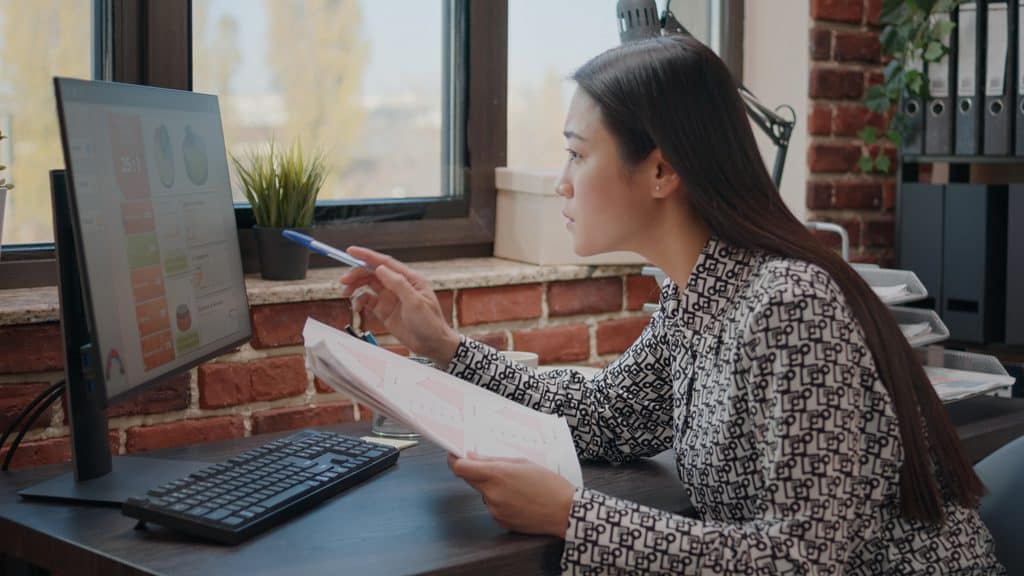 Woman in a patterned blazer holding printed documents while looking at a monitor displaying a colorful business dashboard in a brick-walled office.