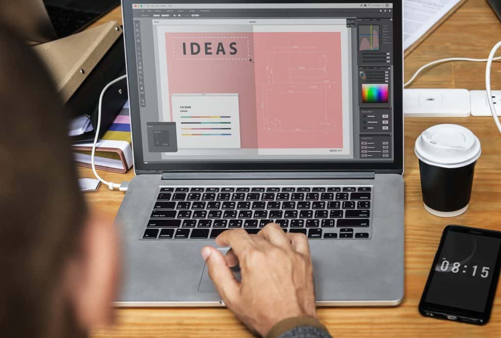 Close-up of a designer's hand on a laptop trackpad while editing a branding and color layout, with a coffee cup on the desk.