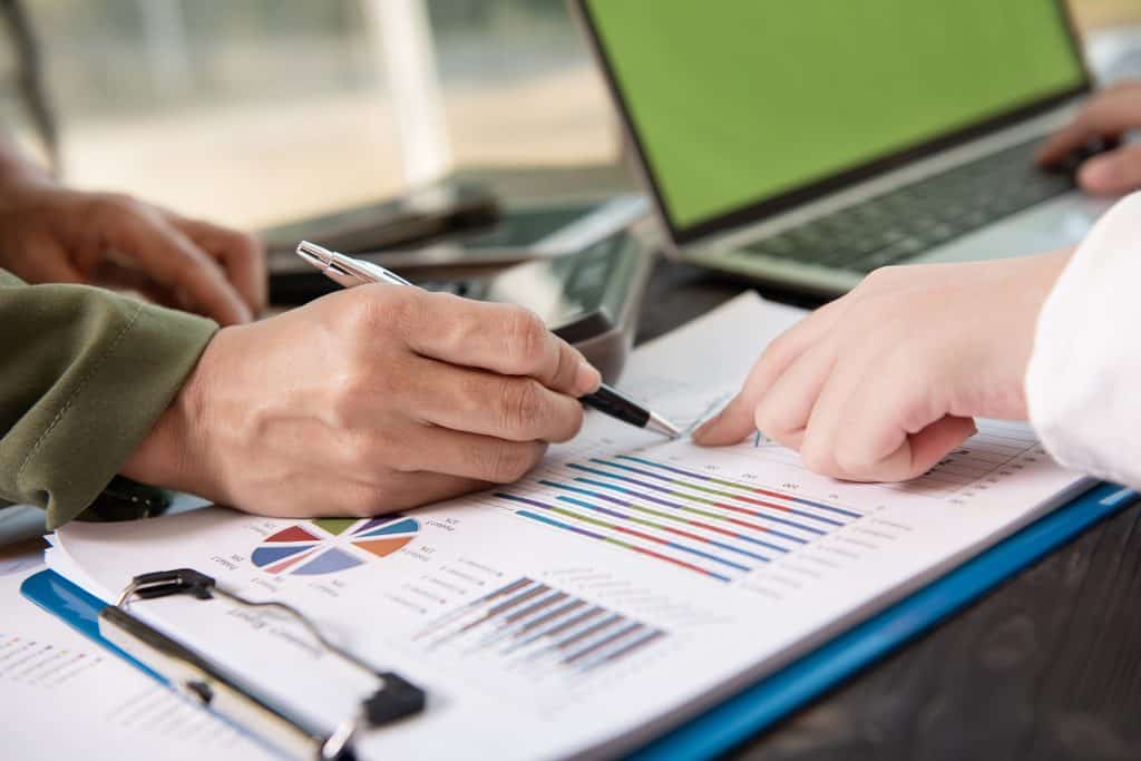 Close-up of two people's hands pointing at printed bar and pie charts on a clipboard, with a pen and open laptop nearby.
