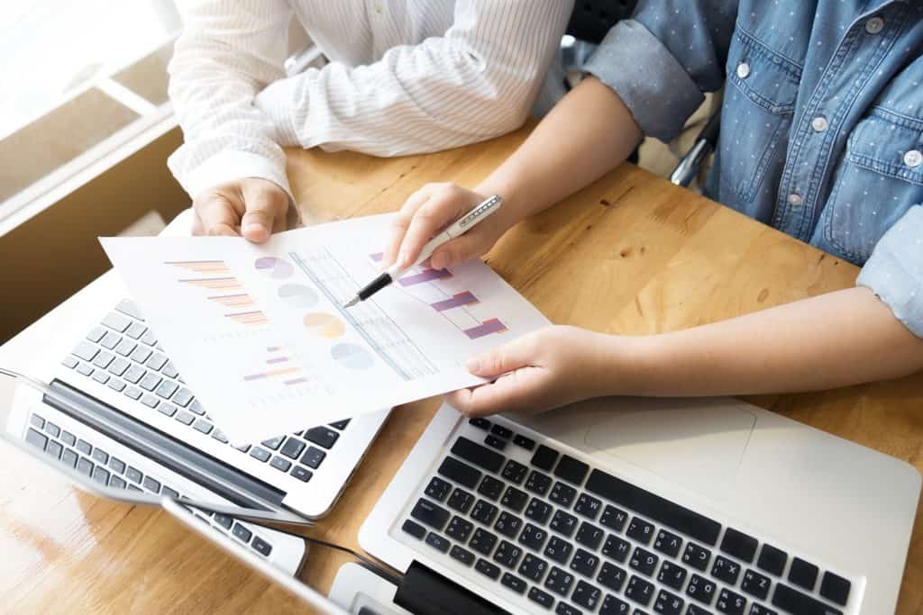 Close-up of two people's hands reviewing a printed bar and pie chart together over two open laptops on a wooden desk.