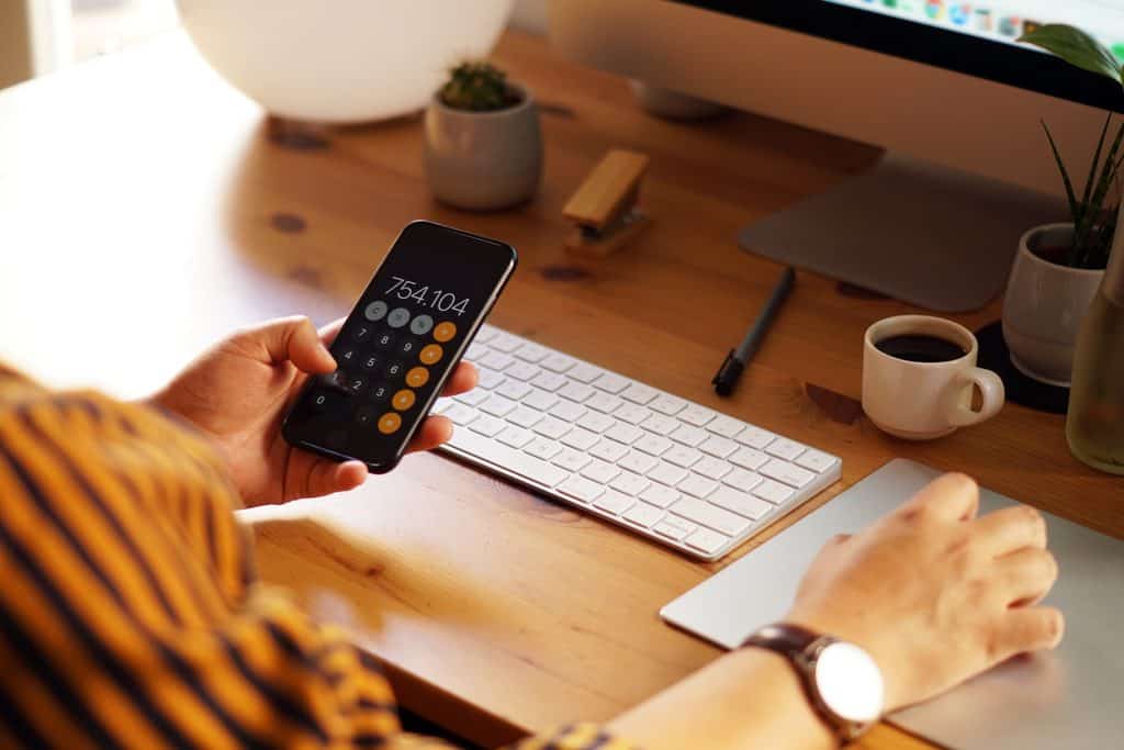 Close-up of a hand holding a smartphone displaying a calculator app with the number 754.104, next to a keyboard and coffee cup on a wooden desk.