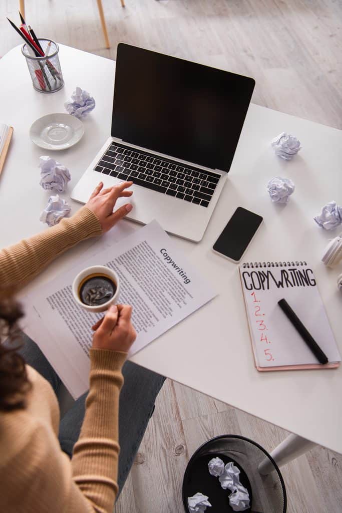 Overhead view of a laptop, coffee cup, crumpled paper, and a notebook labeled "Copywriting" with a numbered list on a desk.