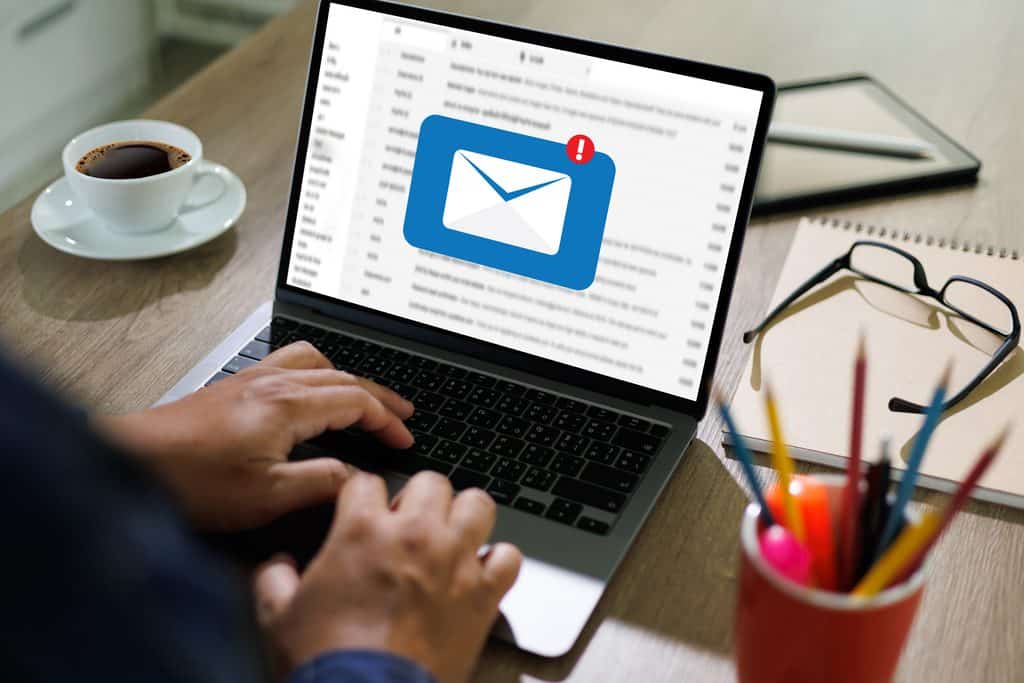 Close-up of hands typing on a laptop showing an email inbox with a large envelope icon and notification badge, coffee and glasses on the desk nearby.