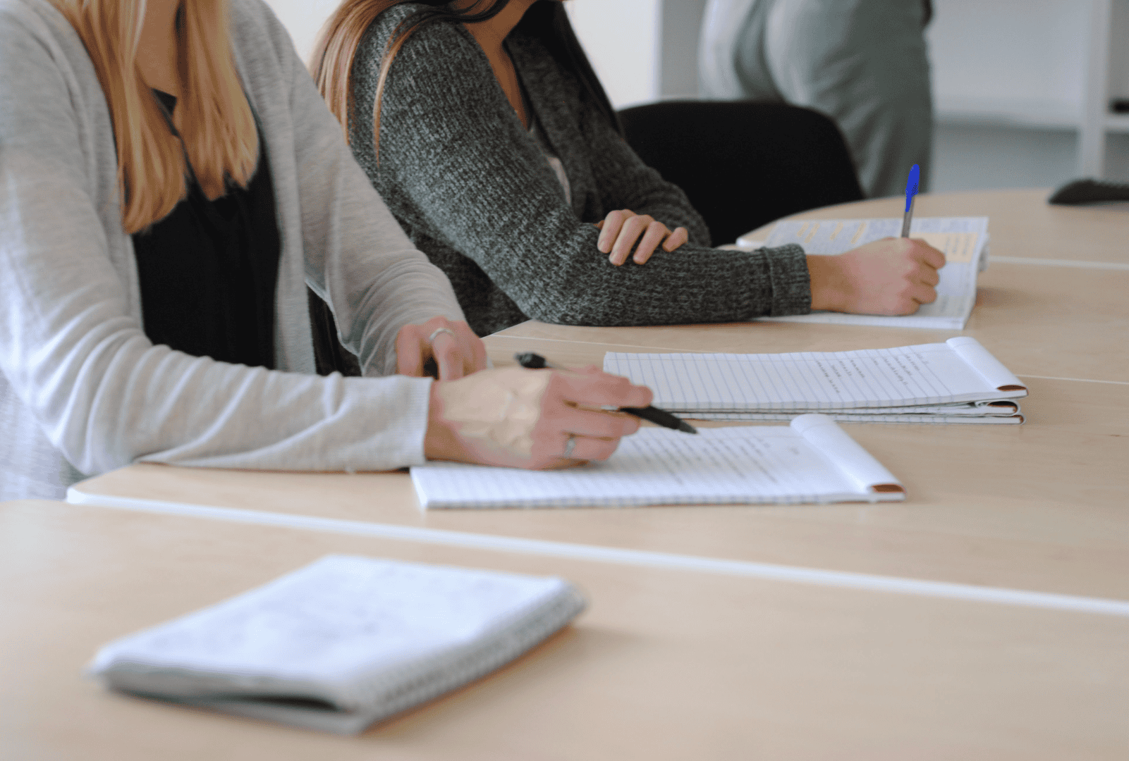 Two students sitting focused at a table, actively taking notes in a notebook with pens.