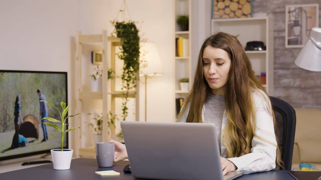 Woman sitting at a desk working on a laptop with coffee nearby, a TV in the background playing an outdoor video clip.