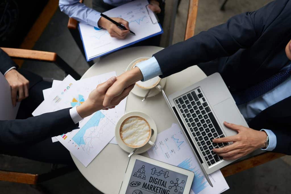 Overhead view of two people shaking hands over a table covered in printed charts, coffee cups, a laptop, and a tablet displaying "Digital Marketing."