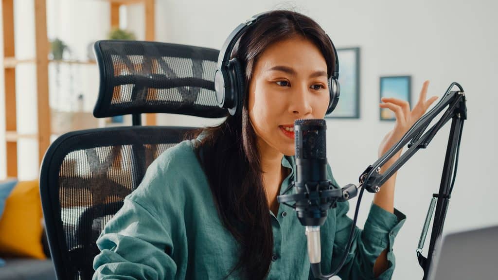 Woman wearing headphones speaking into a studio microphone while gesturing, recording from a home office setup.