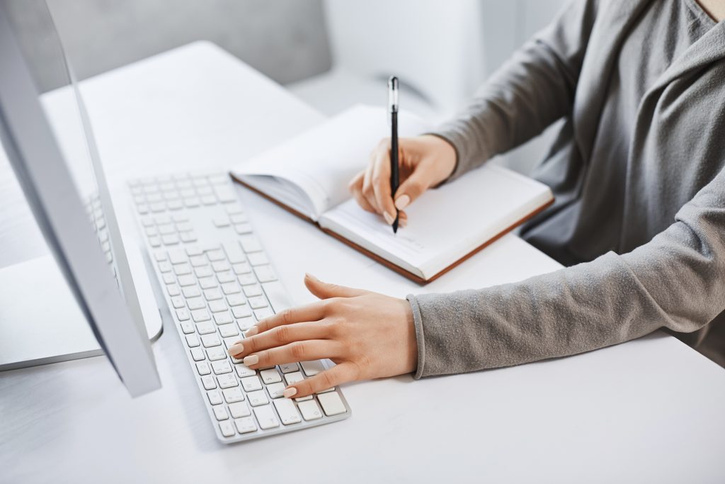 Close-up of a woman's hands typing on a white keyboard with one hand while writing in an open notebook with a pen, beside a desktop monitor.
