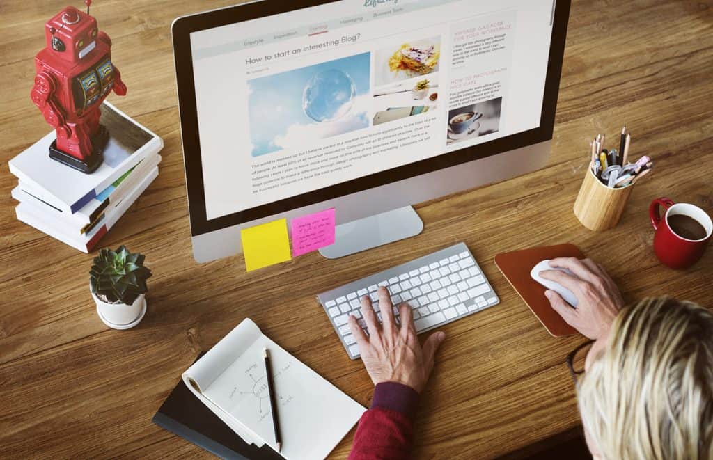 Overhead view of a desktop monitor displaying a blog post titled "How to start an interesting Blog?" beside a handwritten diagram mapping brand strategy, product, team, visual, and customers.