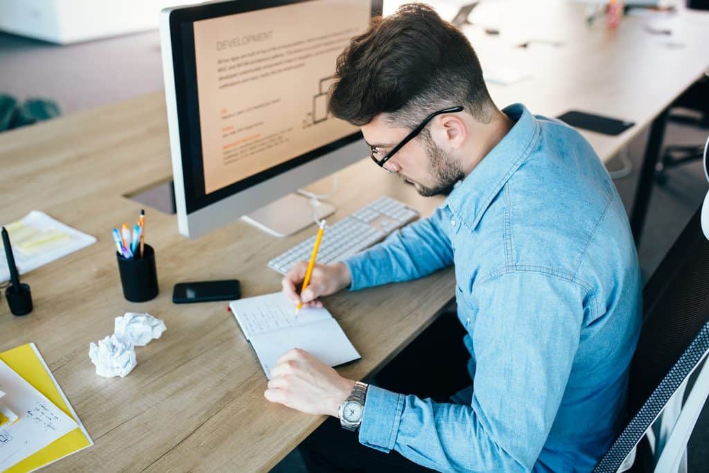 Man in glasses writing notes at a desk beside a monitor displaying development documentation