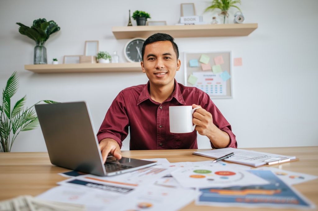 Man in a maroon shirt smiling at the camera while holding a white mug, one hand on a laptop, surrounded by printed charts and reports on a wooden desk.