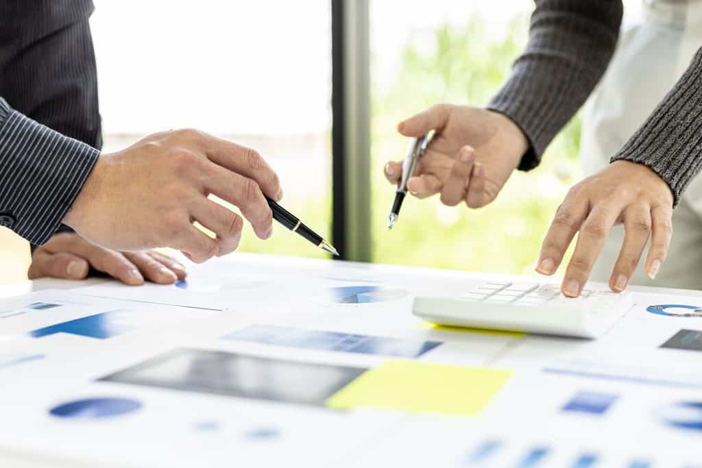 Close-up of two people's hands pointing at printed charts and using a calculator on a desk covered in reports and sticky notes.