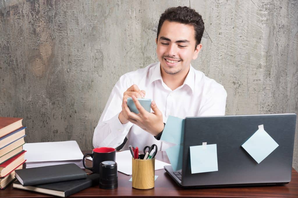 Man in a white shirt smiling while writing on a small sticky note at a desk with books, a laptop, and office supplies.