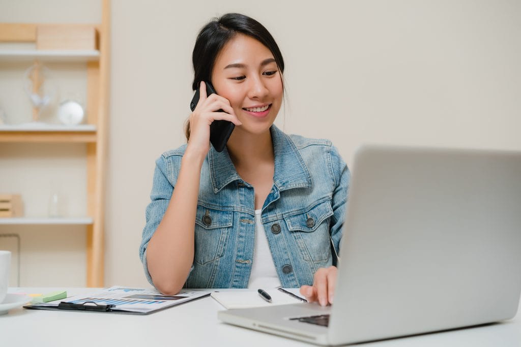 Woman in a denim jacket smiling while talking on the phone and typing on a laptop, with printed charts and a notebook on the desk.