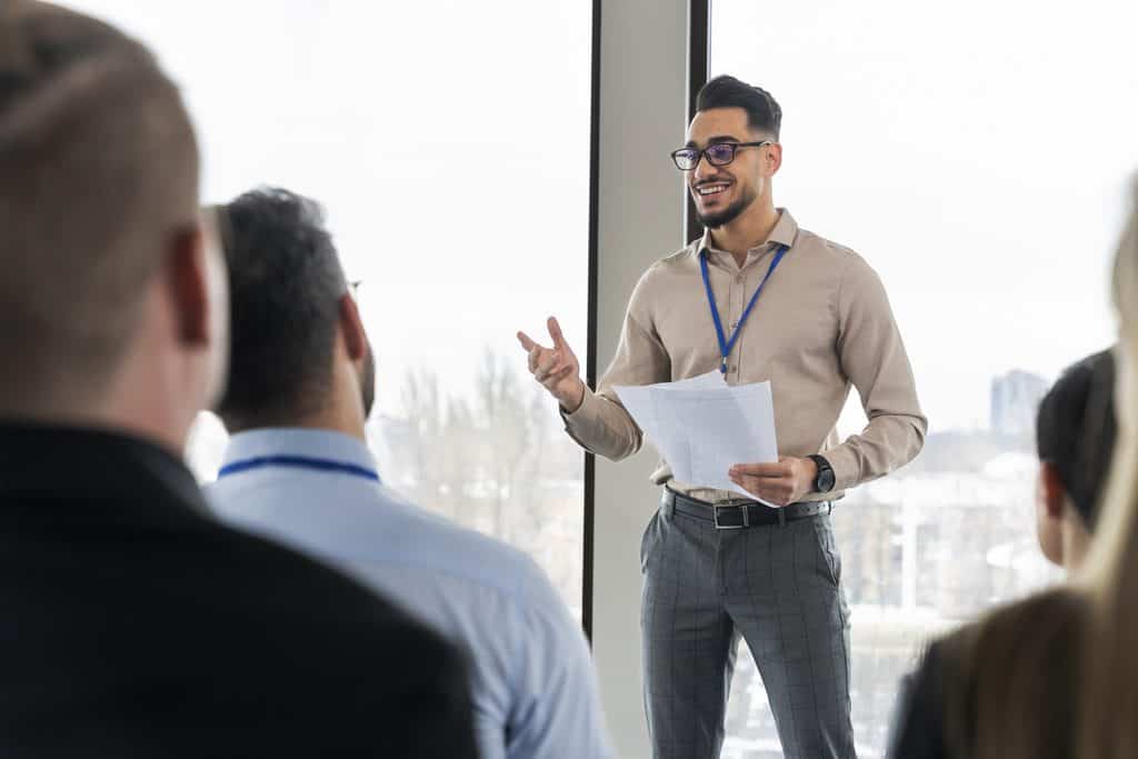 Presenter gesturing while pitching an idea to colleagues in a bright office, an example of how to find ghostwriting clients through in-person networking and agency pitches.