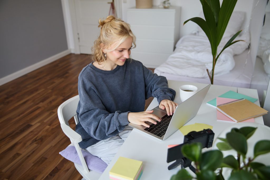 Woman with her hair in a bun smiling while typing on a laptop at a white desk with a camera on a tripod, notebooks, and sticky notes nearby.