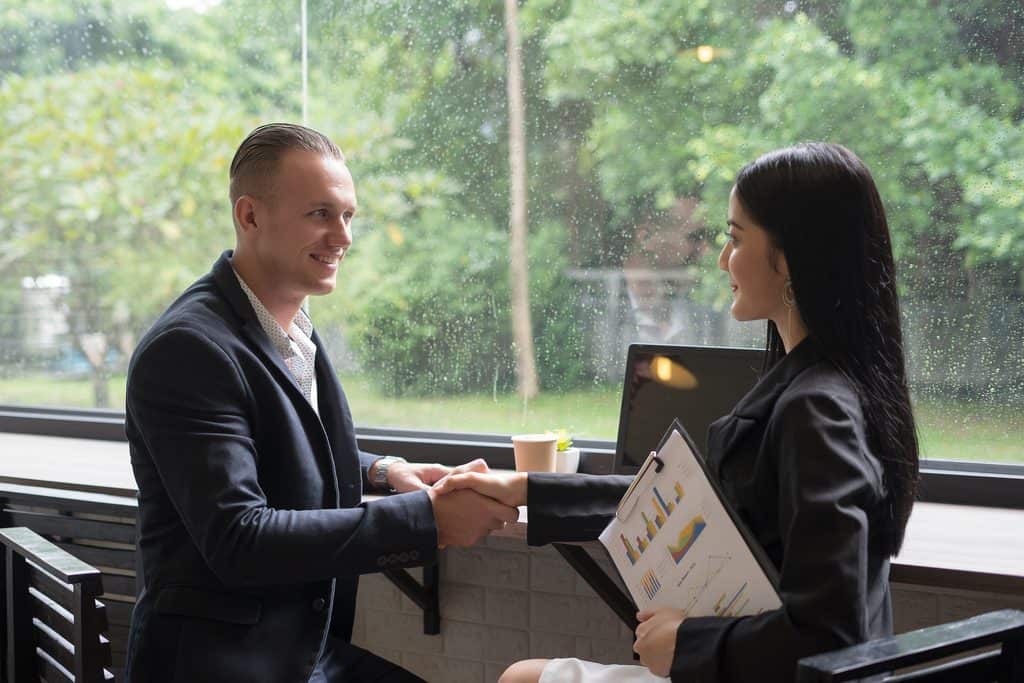 Man and woman shaking hands near a window, with the woman holding a clipboard showing colorful bar and line charts.