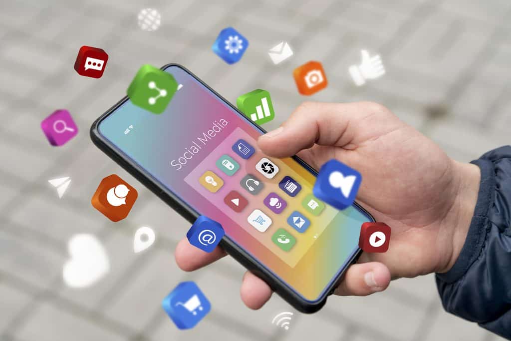 Hand holding a smartphone displaying a "Social Media" screen surrounded by floating icons for messaging, sharing, camera, and analytics apps.