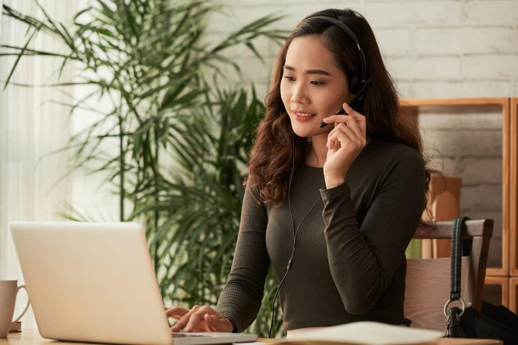 Woman wearing a headset and smiling while working on a laptop at a desk with a coffee mug and a plant nearby.