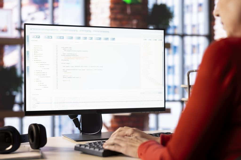 Person in an orange sweater typing at a desk with a large monitor displaying code and a project management interface, headphones nearby.
