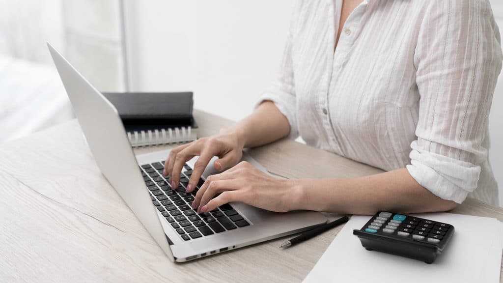 Close-up of a woman's hands typing on a laptop keyboard beside a calculator, notebook, and pen on a desk.