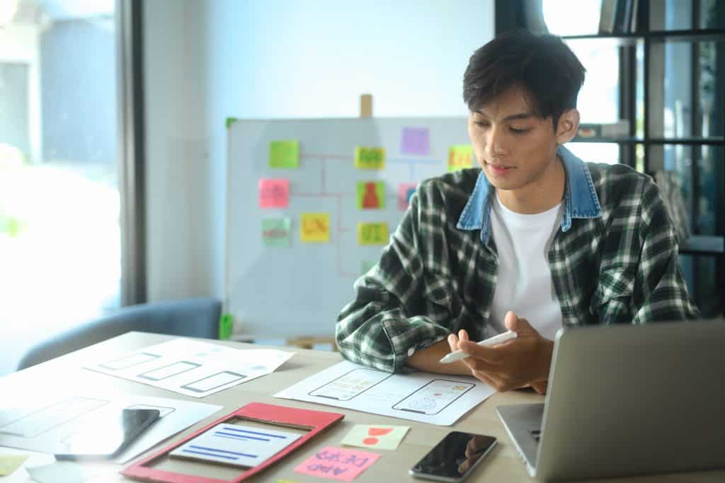 UX designer reviewing hand-drawn wireframes and app sketches spread across a desk while working on a laptop.