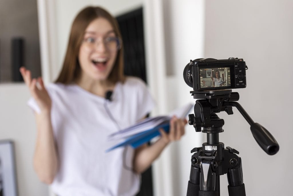 Woman smiling and gesturing while recording herself on a mirrorless camera mounted on a tripod, holding notes.
