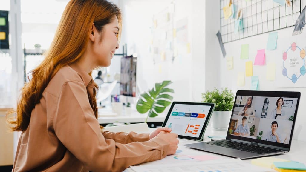 Woman smiling during a video call with four colleagues while reviewing a project roadmap on a tablet at her desk.