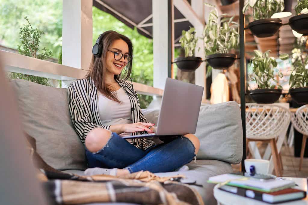 Woman wearing headphones and glasses smiling while working on a laptop on an outdoor couch surrounded by plants.
