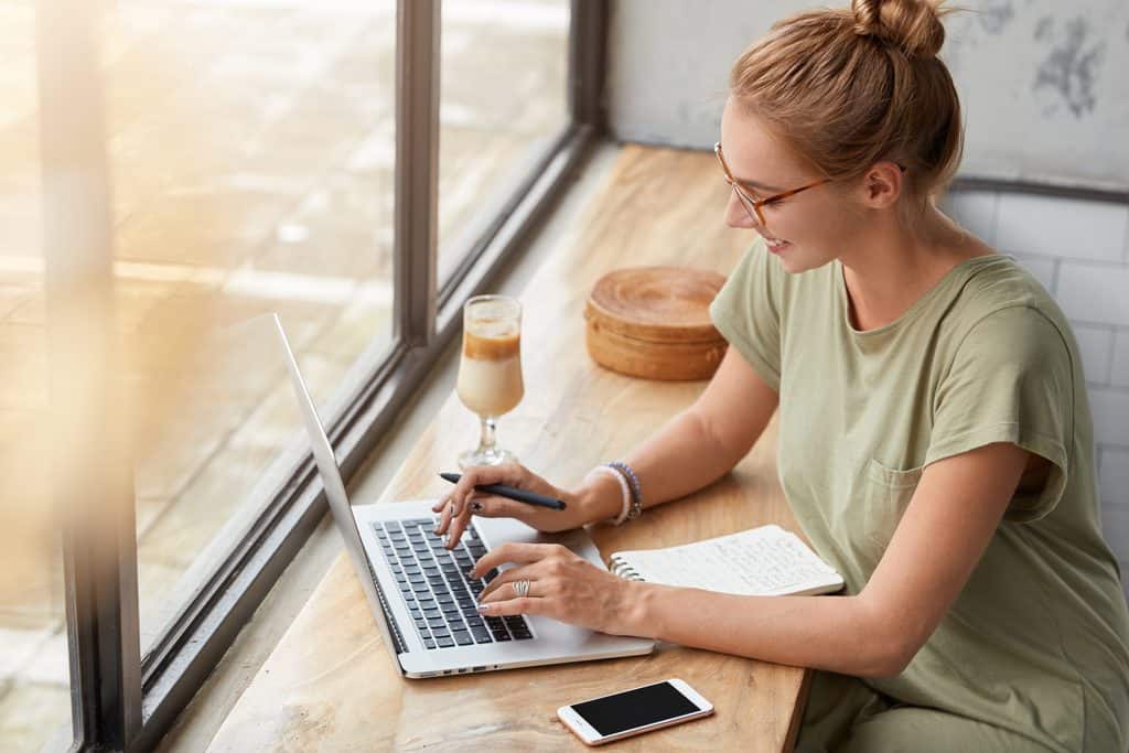 Freelancer writing on a laptop with a notebook at a cafe window counter