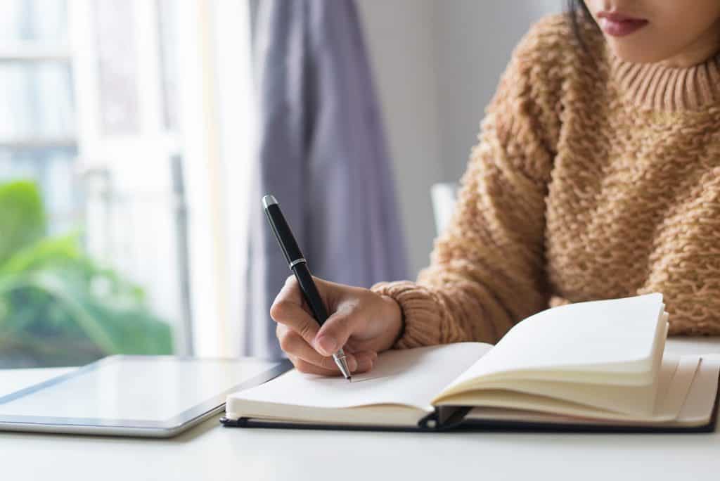 Close-up of a woman's hand writing notes in an open journal beside a tablet on a wooden desk.