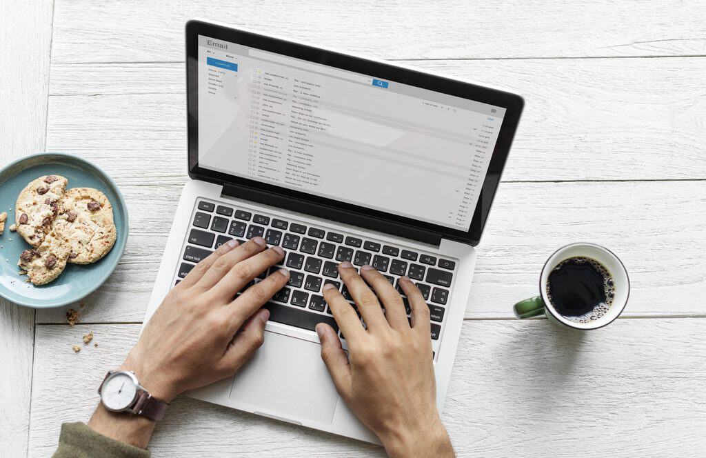 Overhead view of a person’s hands typing on a silver laptop showing an email inbox with a compose button and folders, beside a blue plate of chocolate chip cookies, a cup of black coffee, and a wristwatch on a white wooden table.