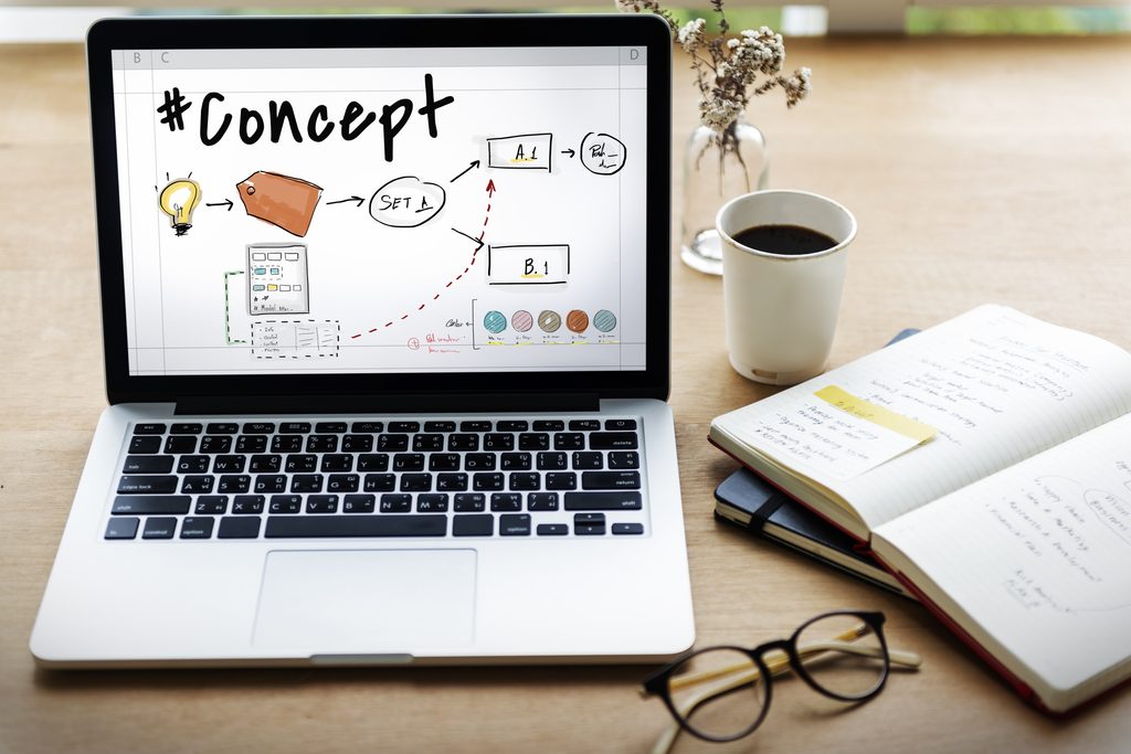 Laptop screen displaying a hand-drawn "#Concept" diagram with a lightbulb, arrows, and labeled boxes, next to a coffee cup, notebook, and glasses on a wooden desk.