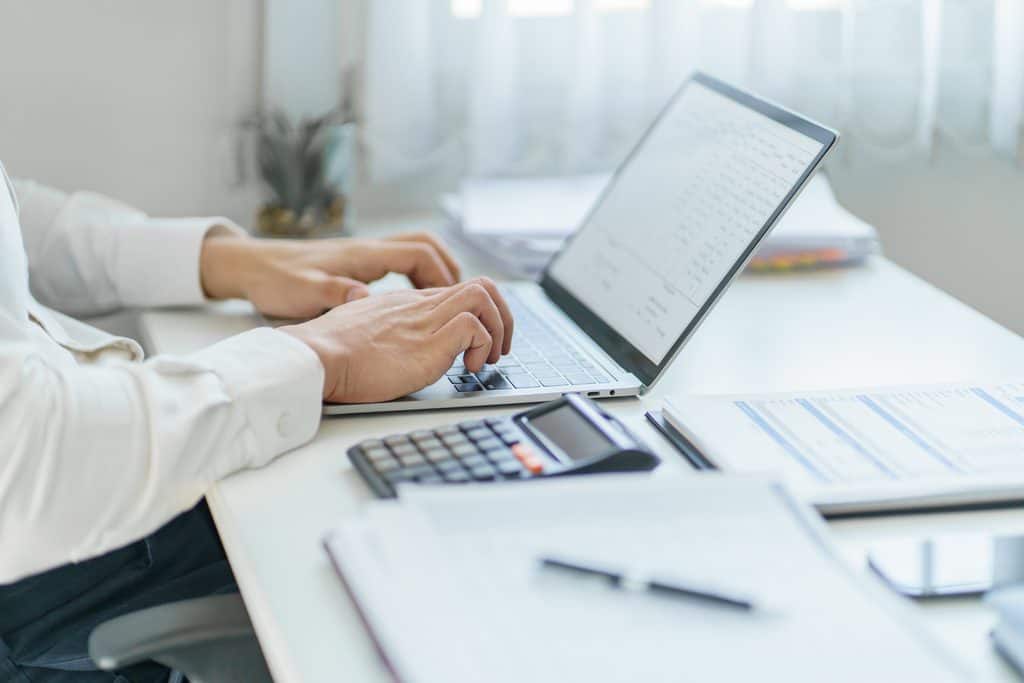 Close-up of a person in a white shirt typing on a silver laptop showing a table, with a calculator, a printed spreadsheet, and stacked papers on a white desk beside a sheer curtain.