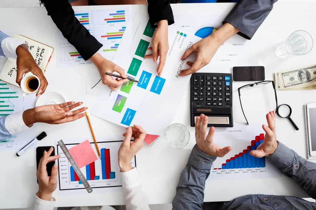Overhead view of several people gathered around a white table covered with bar charts, a diagram of membership tiers, a pie chart, a calculator, a phone, glasses, a magnifying glass, and cash, with hands pointing and gesturing.