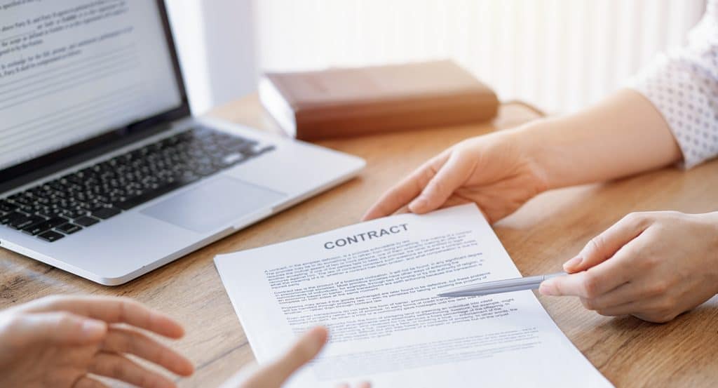 Close-up of two people at a wooden table, one holding a printed document titled Contract while the other points a silver pen at the text, with an open laptop showing a text document and a closed brown notebook nearby.