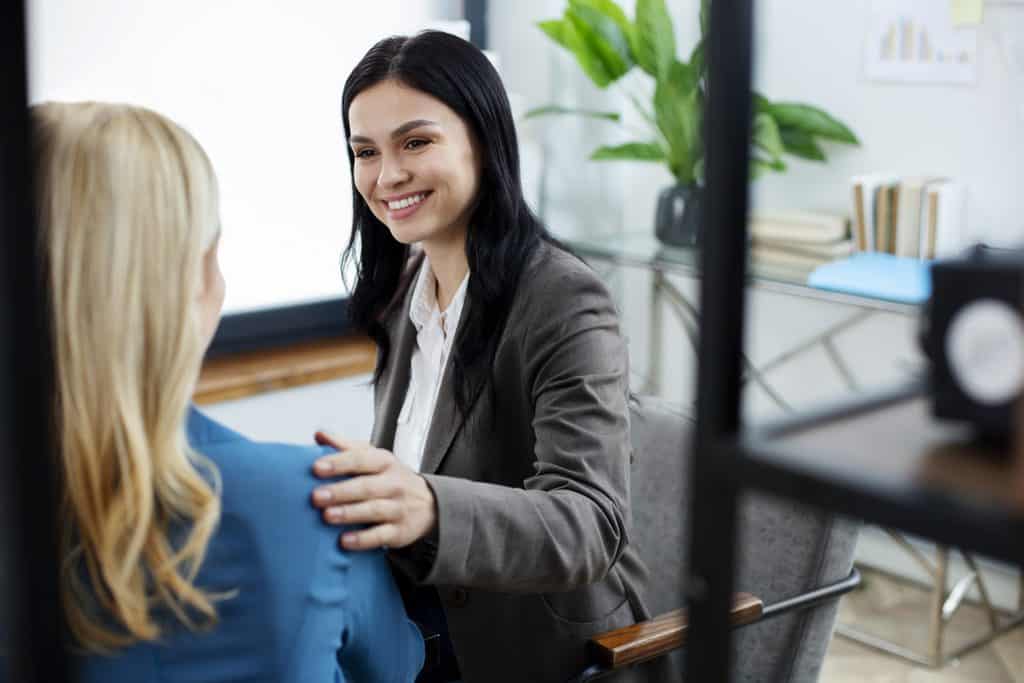 Smiling woman in a gray blazer and white blouse sitting in an office and placing a supportive hand on the shoulder of a blonde woman in a blue top, with a plant and bookshelf in the background.
