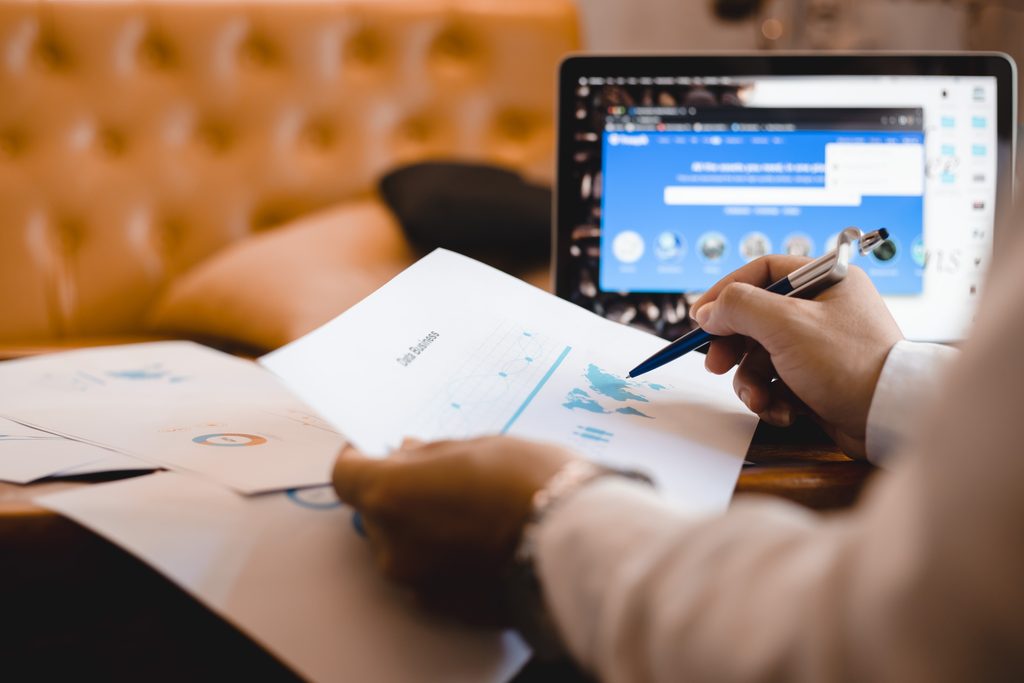 Close-up of a hand holding a pen over a printed "Data Business" report with line charts, a pie chart, and a world map, with a laptop open in the background.